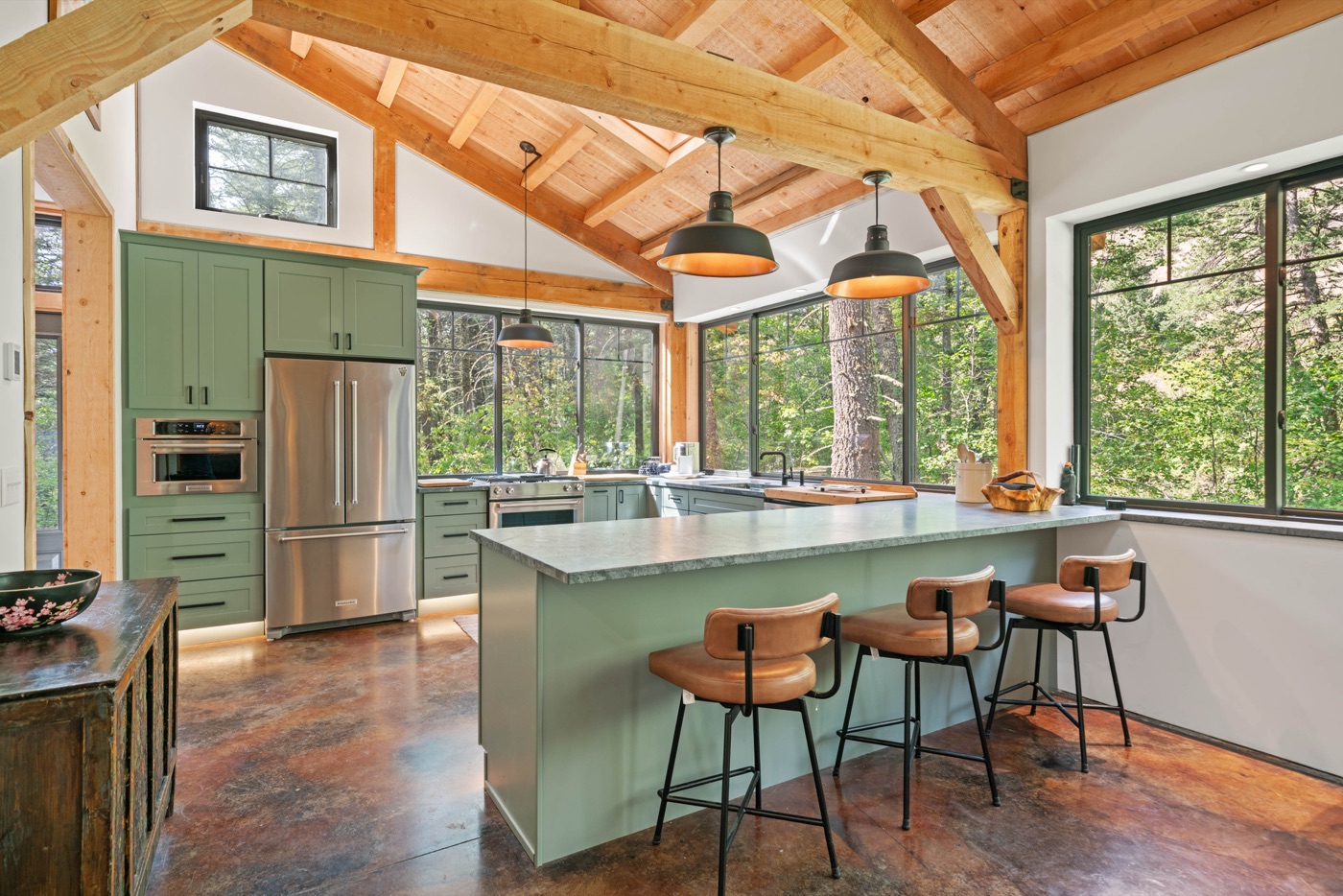Timber-framed kitchen with sage green cabinetry and large windows onto the forest.