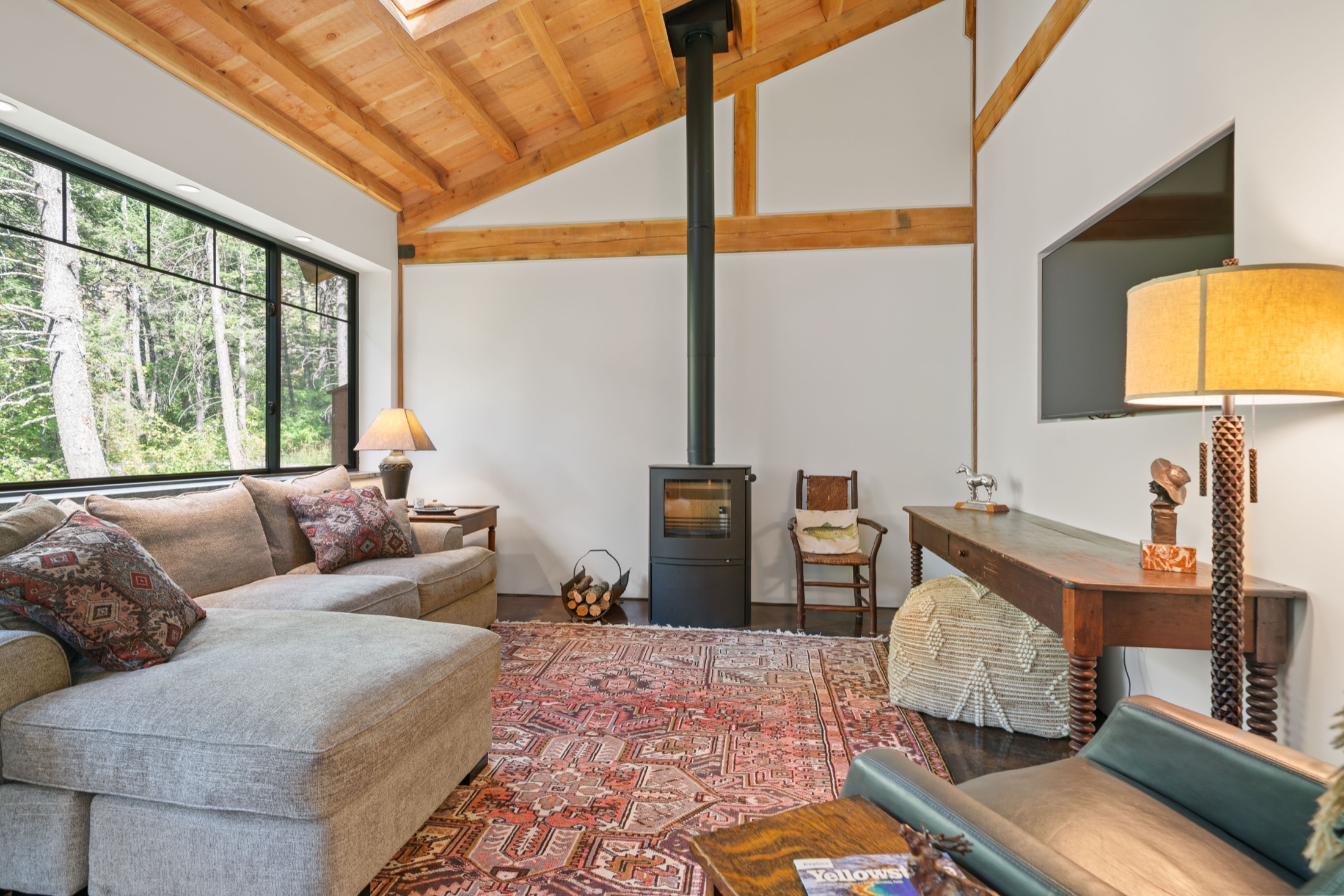 Living room with a vaulted timber ceiling, a wood stove, and a large window onto the trees.